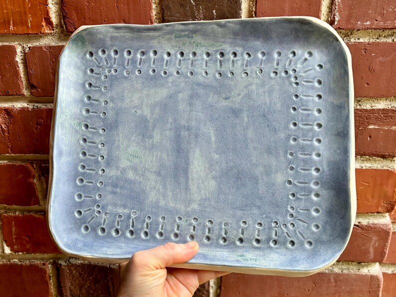Op de afbeelding: Een rechthoekige keramische serveerschaal met een lichtblauwe glazuur. De schaal heeft een decoratieve rand van kleine, gelijkmatig verdeelde gaatjes. De schaal wordt door een hand tegen een bakstenen muur gehouden.