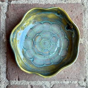 May include: A handmade ceramic bowl with a unique, organic shape. The bowl features a blend of colors, including teal, yellow, and brown, with a textured, honeycomb-like pattern in the center. The rim has a wavy edge.