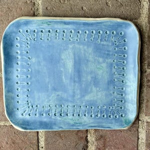 Könnte beinhalten: Ein rechteckiges Keramiktablett mit hellblauer Glasur. Das Tablett hat einen dekorativen Rand aus kleinen, gleichmäßig verteilten Löchern und einen leicht erhöhten Rand. Die Oberfläche weist subtile Farbvariationen auf, was seinen einzigartigen, handwerklichen Reiz unterstreicht.