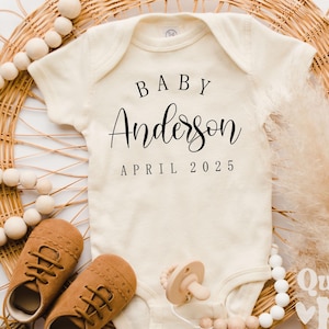 May include: A white baby onesie with the text "BABY Anderson APRIL 2025" in black. The onesie is lying on a woven basket with brown leather baby shoes, a wooden teether, and a white beaded necklace.