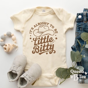 May include: Cream-colored baby onesie with the text "It's Alright to be Little Bitty" in brown. The design includes a cowboy hat and stars. The image also shows baby shoes, a wooden rattle, and denim shorts.