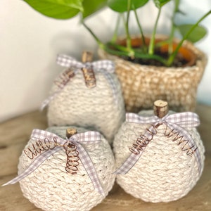 May include: Three crocheted pumpkins in a cream color with a light gray and white gingham ribbon and copper wire accents. The pumpkins are sitting on a wooden surface.