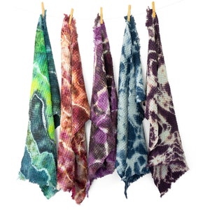 May include: Five tie-dye waffle weave towels hanging from a clothesline. The towels are in shades of green, red, purple, blue, and purple-brown. Each towel has a unique pattern and frayed edges, creating a rustic, handmade aesthetic.