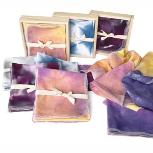 May include: A set of eight tie-dye fabric squares in various shades of purple, blue, and yellow. The squares are folded and tied with twine, and some are displayed in wooden boxes.