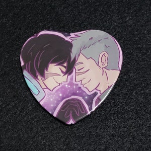May include: Heart-shaped button featuring a cartoon illustration of two men with short hair, one with black hair and the other with gray hair, looking at each other with a galaxy background.