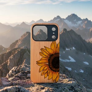 May include: A wooden phone case featuring a detailed illustration of a sunflower. The sunflower is rendered in shades of yellow, orange, and brown, with a dark center. The case has a black camera cutout and is set against a mountain backdrop.