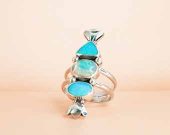 May include: A silver ring featuring three oval and triangular turquoise-colored stones, and one opal-colored stone. The ring has a unique design with multiple bands and decorative accents, set against a soft peach background.