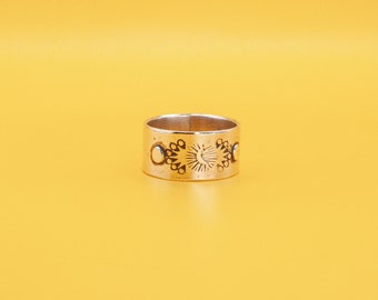 May include: A silver-toned ring with a wide band. The ring features a repeating pattern of sun and crescent moon designs. The ring is set against a yellow background.