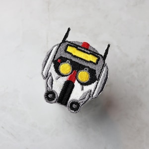May include: Embroidered patch of a white and grey robot head with yellow eyes and a red stripe. The robot has a black visor and a black mouth.