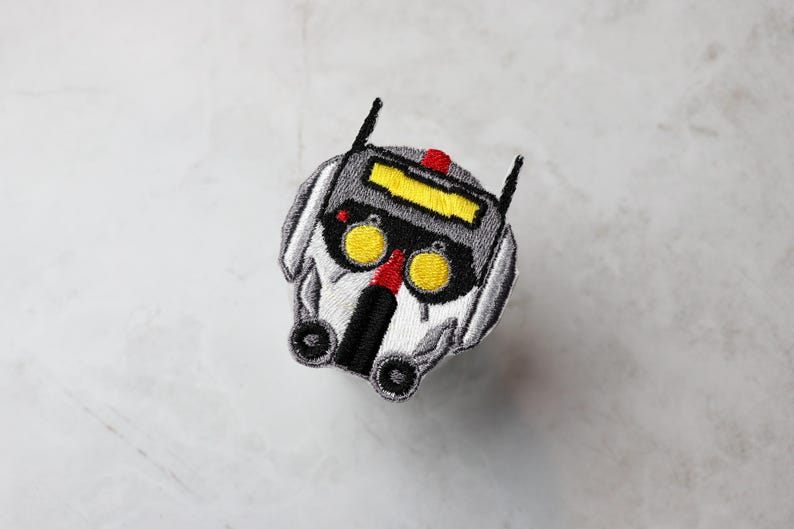May include: Embroidered patch of a white and grey robot head with yellow eyes and a red stripe. The robot has a black visor and a black mouth.