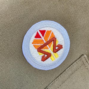 Peut inclure: Un écusson rond blanc avec une bordure bleue. L'écusson présente un motif géométrique dans des tons d'orange, de rouge et de jaune. Le motif ressemble à une chaîne de montagnes stylisée.