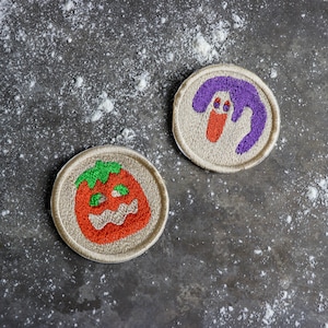 May include: Two embroidered iron-on patches. One patch is a purple ghost with a red tongue. The other patch is a red and green pumpkin with a scary face.