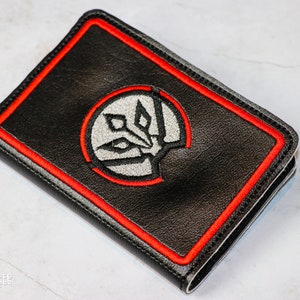 Puede incluir: Cartera negra con borde rojo y un emblema circular. El emblema presenta un diseño blanco y gris. La cartera es rectangular y parece estar hecha de cuero o un material similar. El diseño general es simple y moderno.