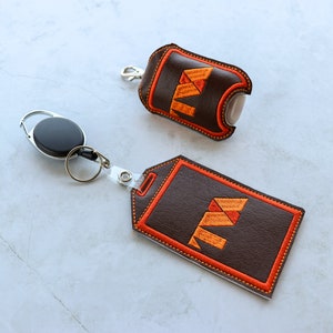 Puede incluir: Un porta-desinfectante de manos de cuero marrón y un porta-insignias de cuero marrón con un carrete de insignia retráctil. Ambos soportes presentan un diseño geométrico naranja y amarillo.