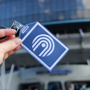 Puede incluir: Un llavero de cuero azul con un logotipo bordado en blanco que presenta un círculo estilizado con un punto en el centro y tres líneas curvas.