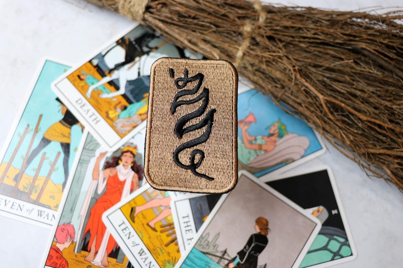 May include: A beige fabric patch with black embroidered text that reads "WITCH". The patch is surrounded by tarot cards, including the Death card, the Ten of Wands card, and the Empress card.