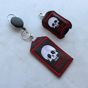 May include: Two black leather keychains with a white skull and the text "22" embroidered on them. One keychain has a retractable cord and the other has a loop.
