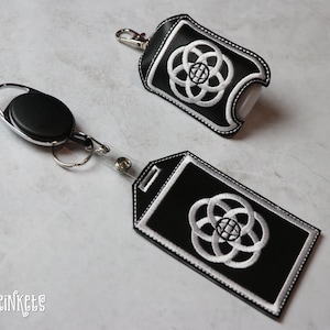 May include: Black leather hand sanitizer holder and a black leather ID badge holder with a white embroidered design of a circle with four connected rings. The holders are attached to a black retractable badge reel.
