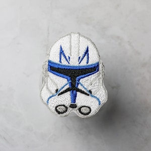 以下が含まれることがあります： 青と黒のアクセントが付いた白いヘルメットの刺繍パッチ。デザインは、SFやファンタジーの世界観からインスピレーションを得た、様式化されたヘルメットを描いています。このパッチは、衣類やアクセサリーへの適用を目的としています。