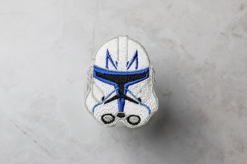 May include: Embroidered patch featuring a white helmet with blue and black accents. The design depicts a stylized helmet with a distinctive shape, likely from a science fiction or fantasy theme. The patch is likely for clothing or accessories.