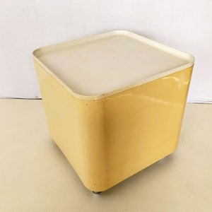 Peut inclure: Un pouf de rangement en plastique jaune et blanc avec un dessus carré et des bords arrondis. Le pouf est doté de quatre petites roulettes.