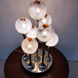 May include: A unique table lamp with multiple frosted glass globes, each with a small orange accent. The globes are supported by chrome stems and arranged on a circular chrome base. The lamp is lit, emitting a warm glow.