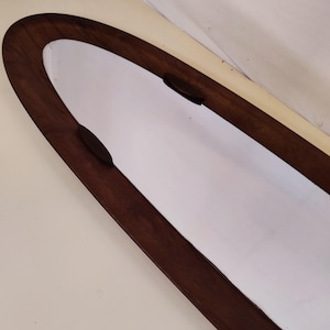 May include: A large oval mirror with a dark brown wooden frame. The frame has a slightly rounded edge and is attached to the wall with two small black brackets.