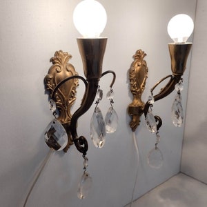 May include: A pair of antique gold wall sconces with crystal drops. Each sconce has a decorative scroll design and a single light bulb.
