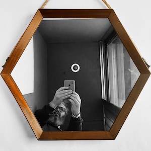 May include: A man is reflected in a hexagonal mirror with a wooden frame. He is holding a smartphone in his right hand and taking a selfie.