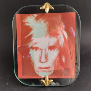 May include: A glass picture frame with a gold-colored metal trim and a decorative fleur-de-lis design on each corner. The frame holds a red and white photograph of a man with a white, spiky hairstyle.