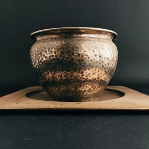 May include: A hammered metal planter with a wide, rounded body and a slightly flared rim. The planter sits on a square, shallow tray. The metal has a mottled, aged appearance with shades of brown and silver.