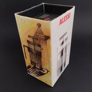 May include: A white box with the word "ALESSI" printed in red on the front. The box features a black and white illustration of a coffee press on the front and a yellow and white illustration of a coffee press on the side.
