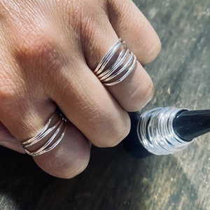 May include: A set of silver stacking rings. The rings are thin and have a hammered texture. The rings are worn on a finger and a stack of rings is shown on a table.