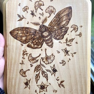 May include: A wood-burned art piece featuring a large moth with detailed wing patterns, surrounded by stylized leaves and floral elements. The artwork is on a light-colored wooden plaque with a unique octagonal shape. The artist's signature is visible.