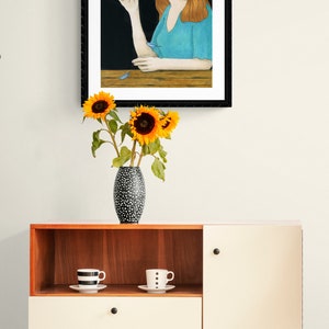 May include: A framed illustration of a woman with long red hair sitting at a table with a blue bird perched on her finger. The background is black and the woman is wearing a light blue shirt.