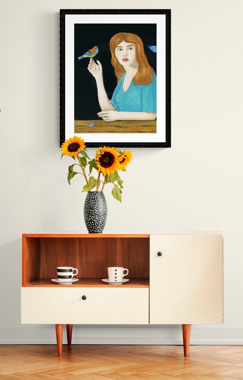 May include: A framed illustration of a woman with long red hair sitting at a table with a blue bird perched on her finger. The background is black and the woman is wearing a light blue shirt.