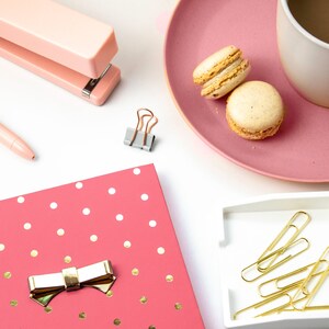 May include: A pink polka dot notebook with a gold bow, a pink stapler, a cup of coffee, two macarons, and gold paper clips in a white dish.