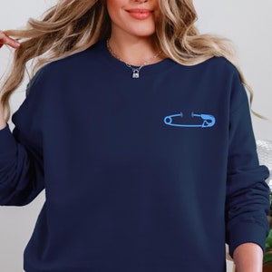 Puede incluir: Una sudadera azul marino con un gráfico de un alfiler de seguridad azul claro en la parte delantera.