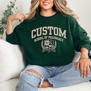 May include: Green sweatshirt with the text "CUSTOM SCHOOL OF PHARMACY" and a crest with a mortarboard, a snake, a pill bottle, and the text "CLASS OF 2024".