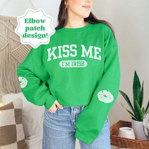 Puede incluir: Sudadera verde con el texto "Kiss Me I'm Irish" en blanco. La sudadera tiene una impresión de labios blancos en cada codo.