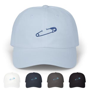 Puede incluir: Un conjunto de cinco gorras de béisbol de diferentes colores, cada una con un diseño de alfiler de seguridad bordado en azul. Las gorras están disponibles en azul claro, blanco, azul marino, marrón oscuro y negro.