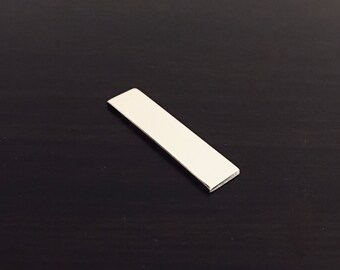 Op de afbeelding: Een klein, rechthoekig object van zilverkleurig metaal. Het object ligt diagonaal op een donkere ondergrond. Het lijkt een kleine staaf of plaat te zijn, met een schoon, reflecterend oppervlak.