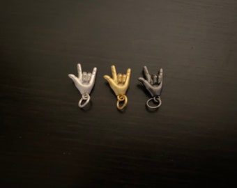 Puede incluir: Tres abalorios plateados en forma de mano haciendo el signo de la lengua de señas americana para 'Te amo'. Los abalorios son de plata, oro y negro.