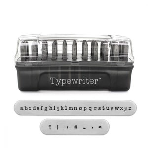 May include: A black plastic case with a clear lid containing a set of metal letter stamps. The case has the word "Typewriter" printed on it. The stamps are arranged in rows and include letters, numbers, and punctuation marks. There are two separate metal strips with the letters of the alphabet and punctuation marks.