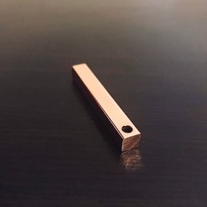 Puede incluir: Una barra de metal de color oro rosa con un agujero redondo en el centro. La barra es de forma rectangular y tiene un acabado liso y pulido.