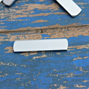 May include: A silver metal bar with rounded edges. The bar is lying on a blue and brown wooden surface.