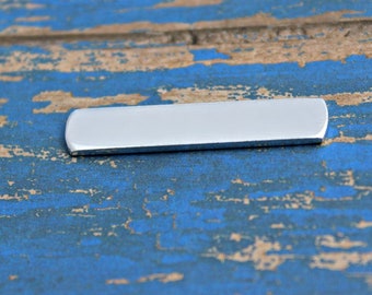 Op de afbeelding: Een zilveren metalen staaf met afgeronde randen. De staaf ligt op een blauw en bruin houten oppervlak.