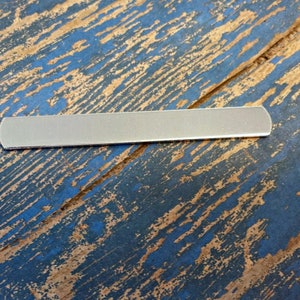 May include: A silver metal nail file with rounded edges, lying on a blue and brown wooden surface.