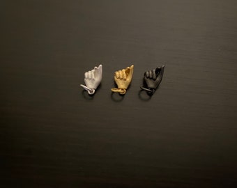 Puede incluir: Tres pequeños amuletos de mano en plata, oro y negro. Cada amuleto tiene forma de mano haciendo el signo shaka.