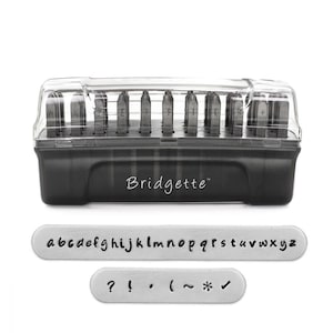 May include: A black plastic case with a clear lid containing a set of metal letter stamps. The case has the word "Bridgette" printed on it. The stamps are arranged in rows and include the alphabet, numbers, and punctuation marks. The stamps are shown on a white background.